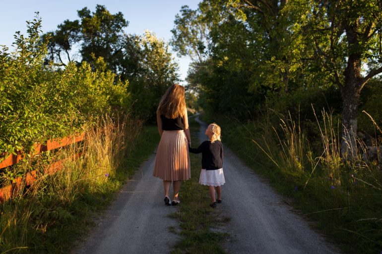 mother and daughter walking on gravel road surrounding by trees and brush